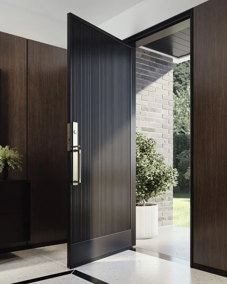 Ribbed matte-black pivot front door with brass hardware beside brick and topiary