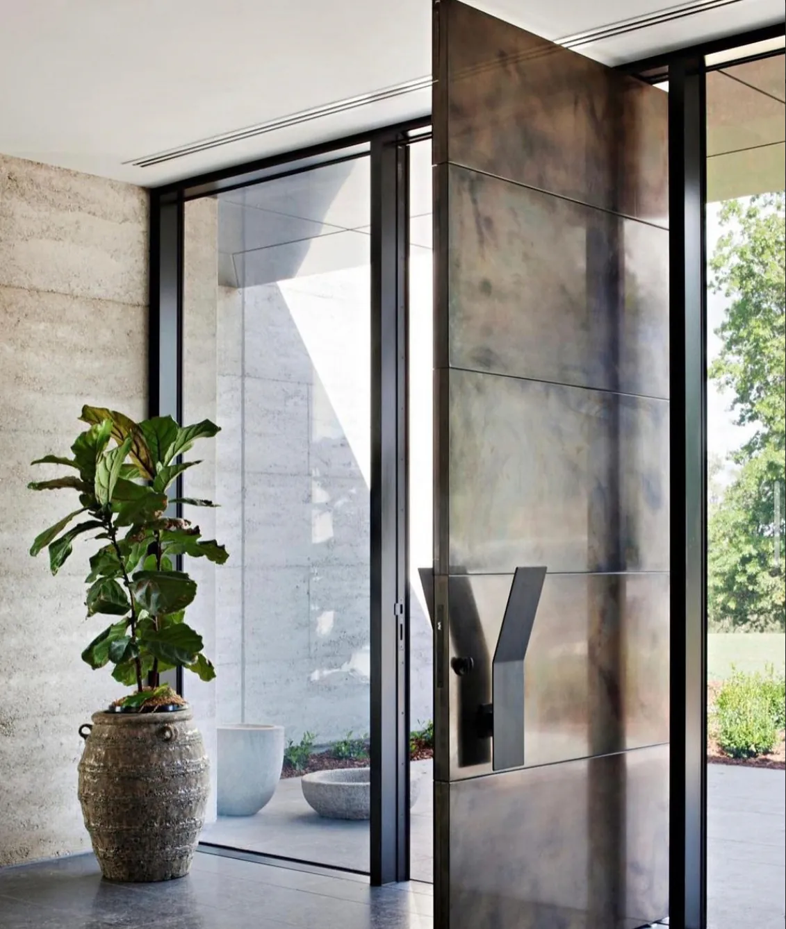 Patinated bronze pivot front door with a sculptural handle beside a glazed entry