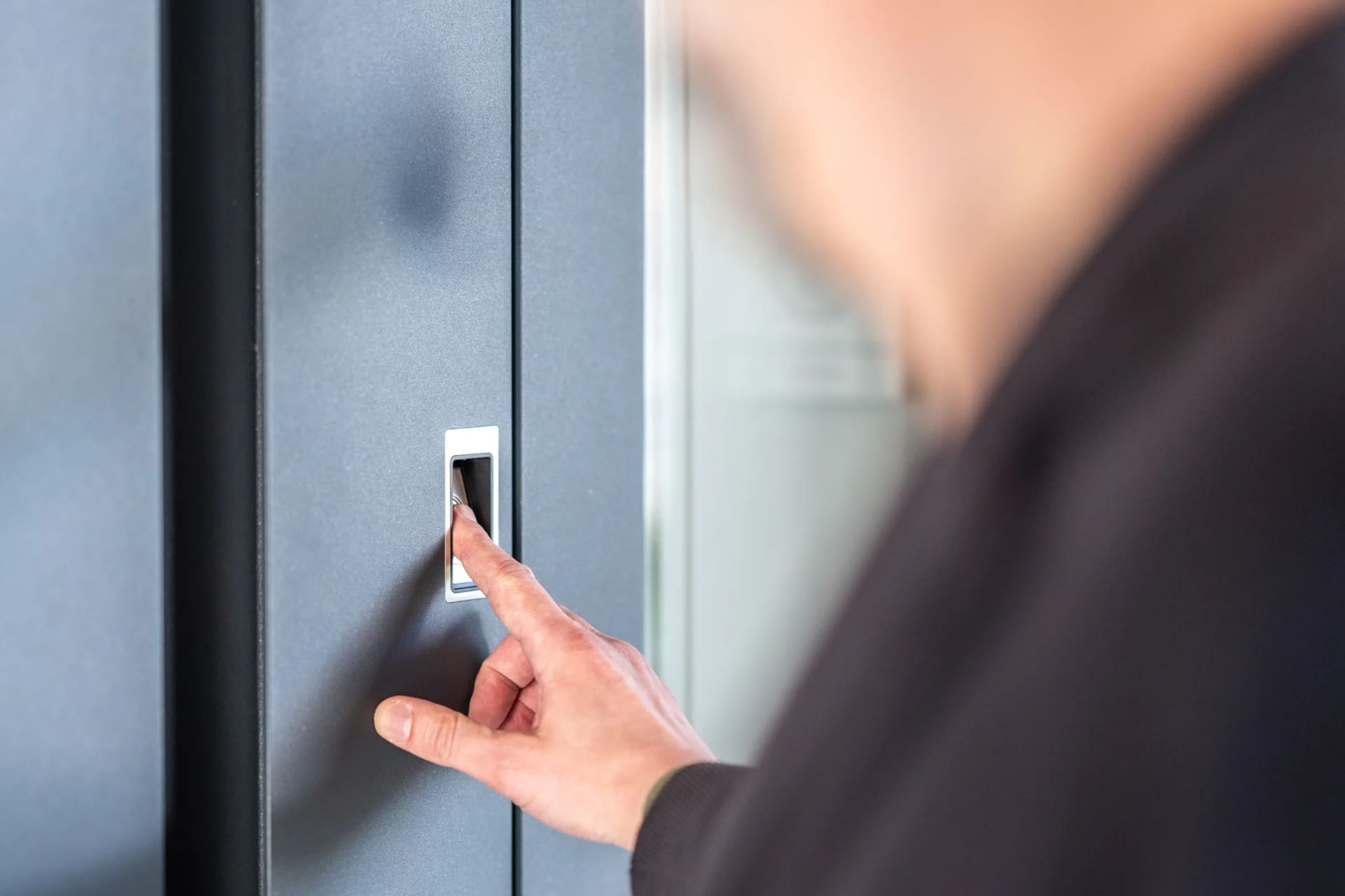 A finger pressing a brushed-steel fingerprint reader set flush into an anthracite front door